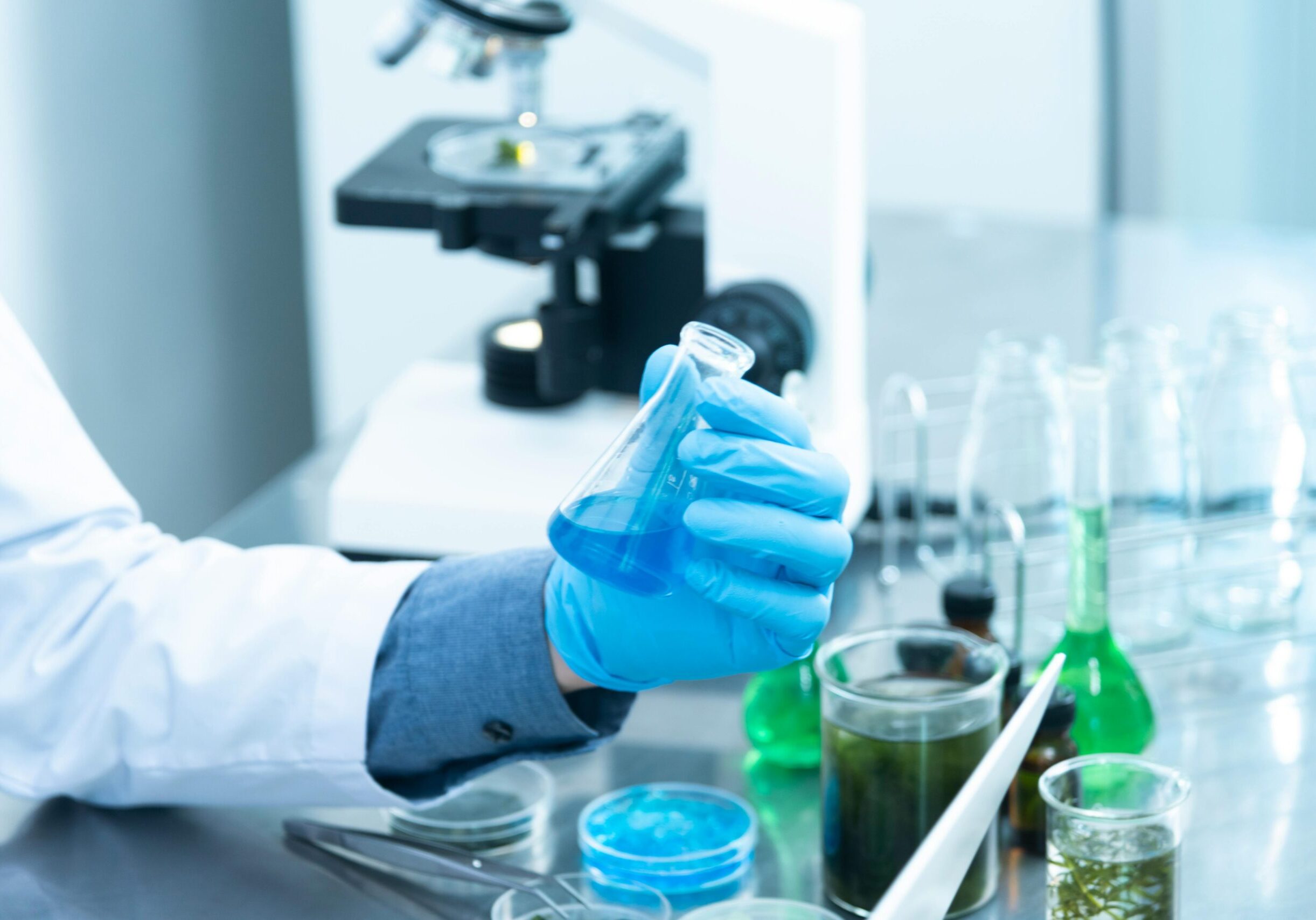 Scientist in gloves analyzing blue liquid in a laboratory setting with microscope and glassware.